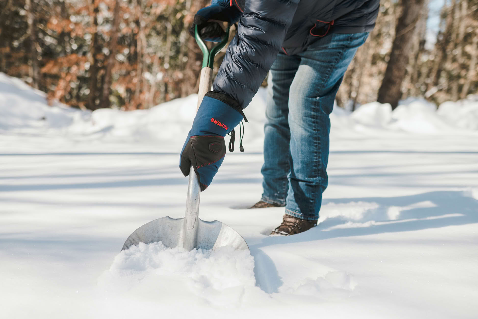 Person wearing gloves and a jacket shovels snow in a snowy outdoor setting.