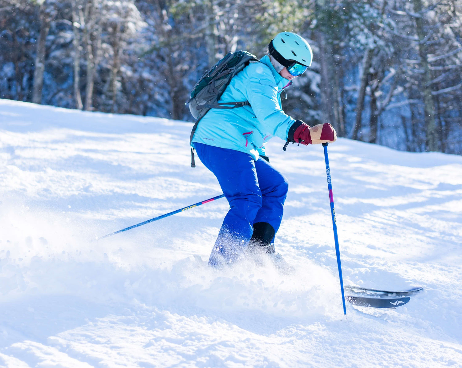 Person skiing downhill on a snow-covered slope, wearing a light blue jacket, blue pants, helmet, goggles, and carrying a backpack.