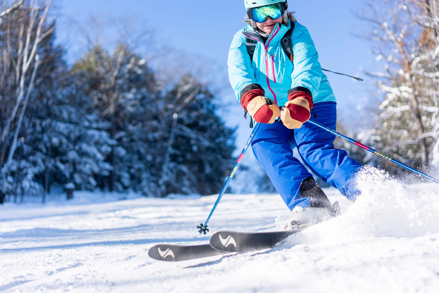 Person skiing downhill on a snowy slope, wearing a blue jacket, blue pants, and goggles, with trees in the background.
