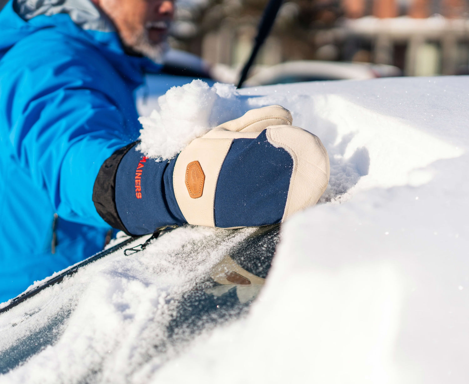 Person in blue jacket clearing snow from a car windshield using beige-and-blue mittens.