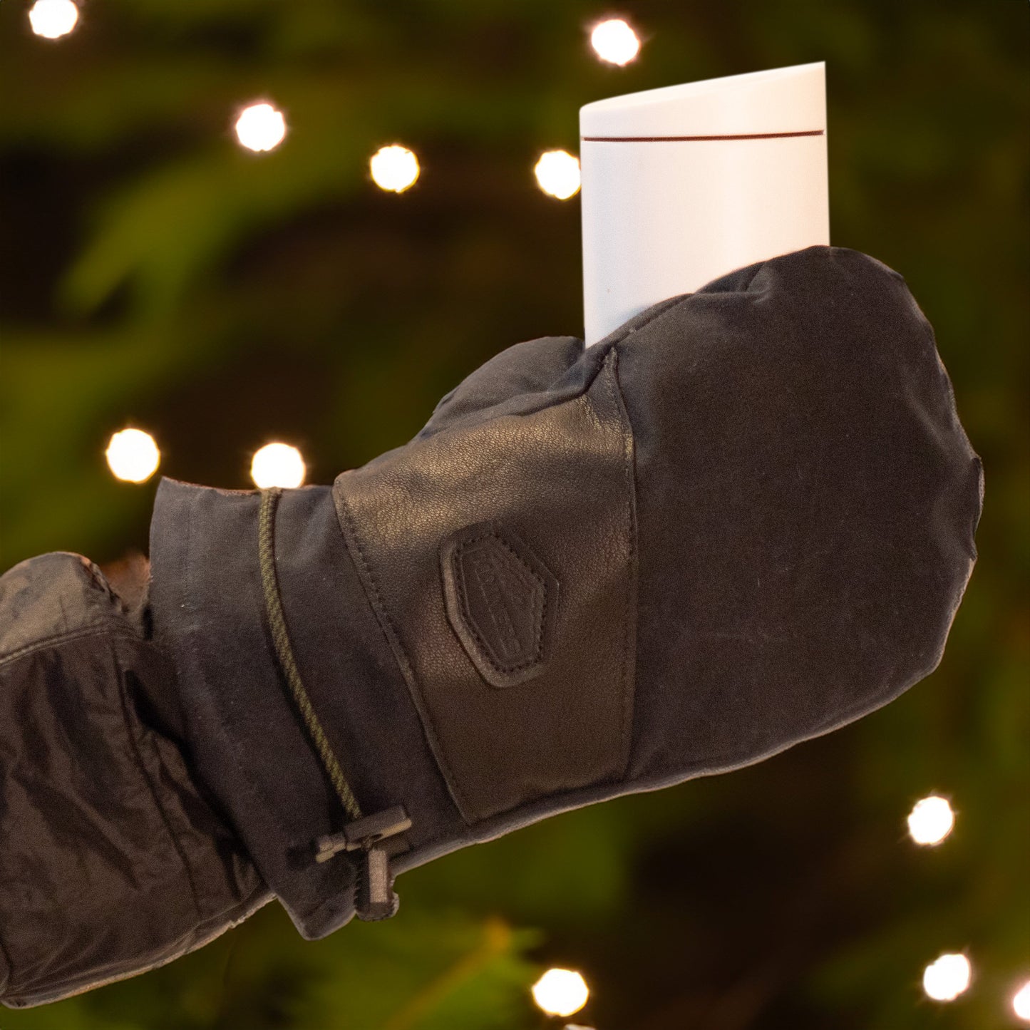 A hand wearing a chic Mainers' Portland Mitt in Black - Discontinued holds a small white gift in front of blurred holiday lights.