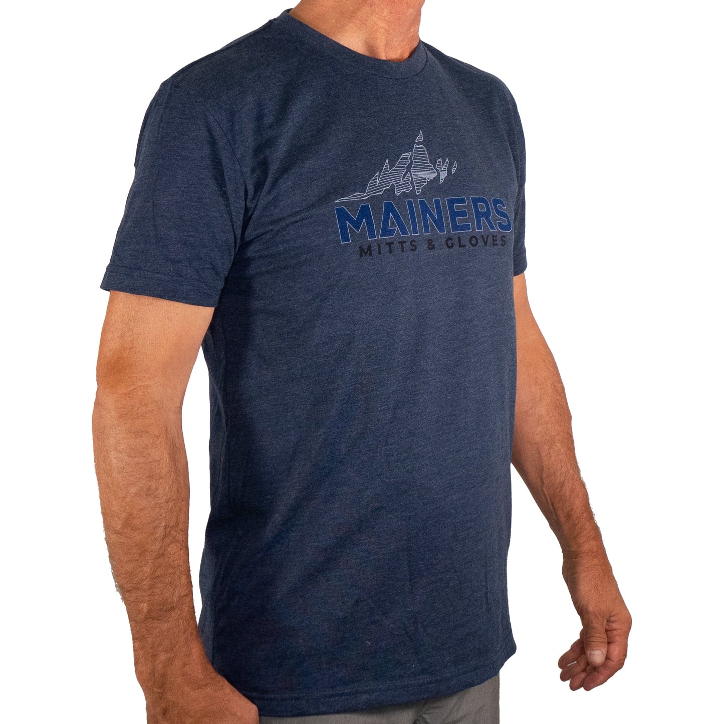 A person sporting a Mainers MG Tee in navy blue, showcasing the "Mainers Mitts & Gloves" slogan and a mountain design, is standing with arms at their sides, poised for an adventurous outdoor journey.
