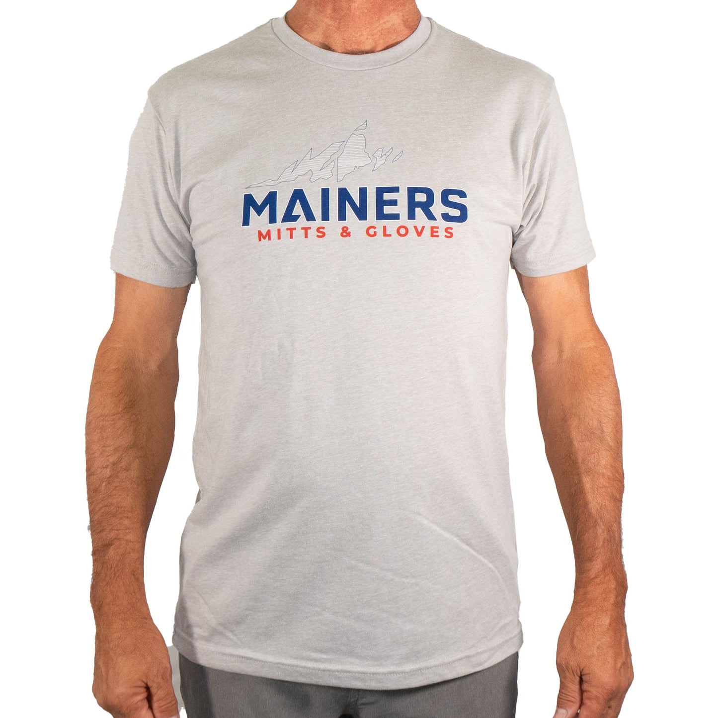 A person is wearing a Mainers MG Tee in light gray, showcasing a mountain graphic with the words "MAINERS MITTS & GLOVES" in blue and red, ideal for outdoor adventures.