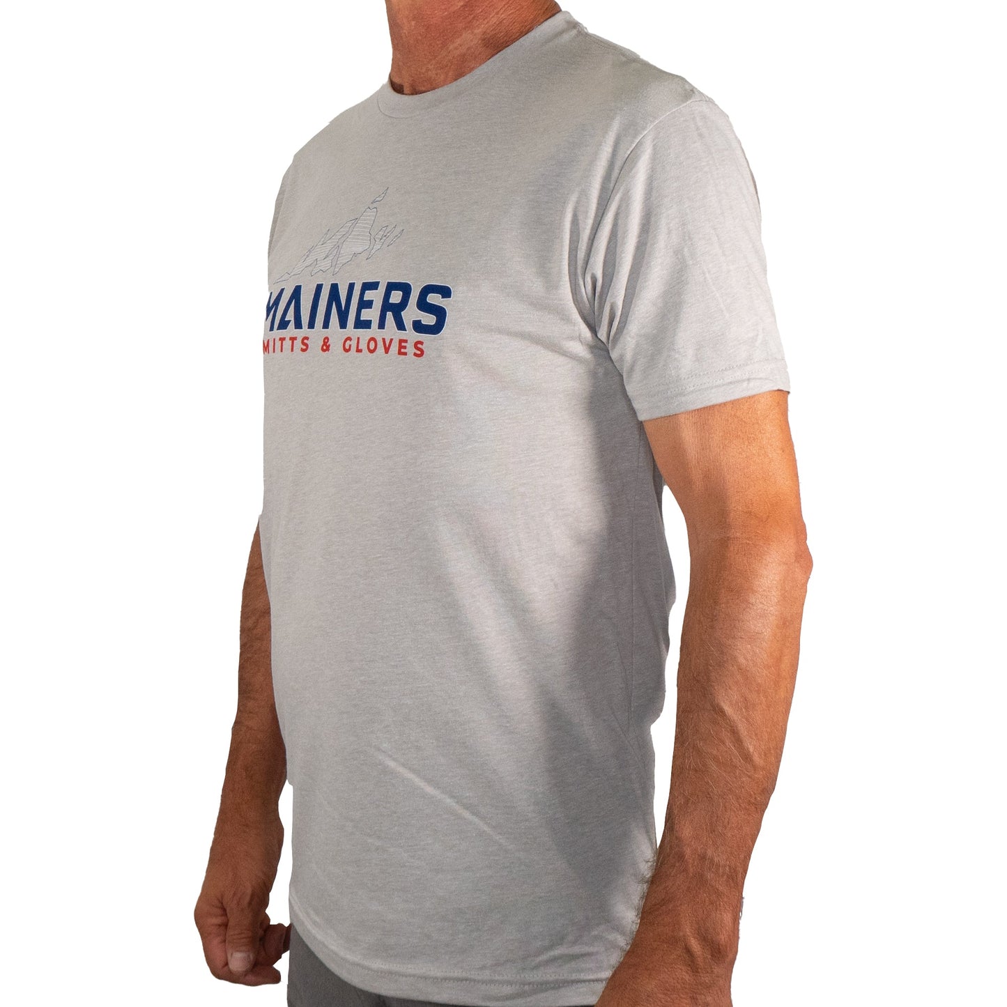 An adventurous individual is captured in a side profile, sporting a gray Mainers MG Tee from the Mainers brand, featuring bold "MITTS & GLOVES" text in striking blue and red. Ideal for those who love outdoor exploration.