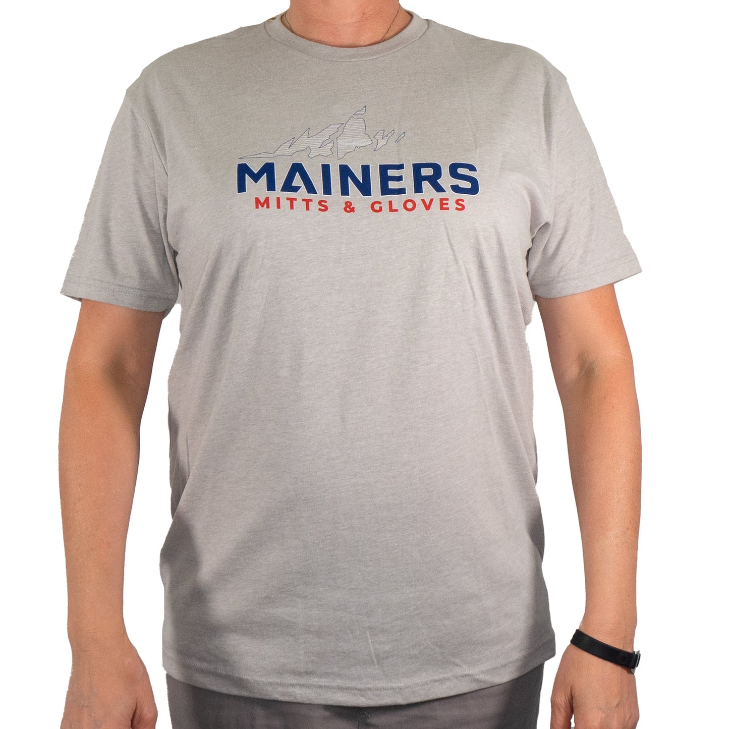 A person donning the Mainers MG Tee, characterized by its gray color and featuring the text "MAINERS MITTS & GLOVES" alongside a small graphic, exudes an adventurous spirit ideal for outdoor exploration.