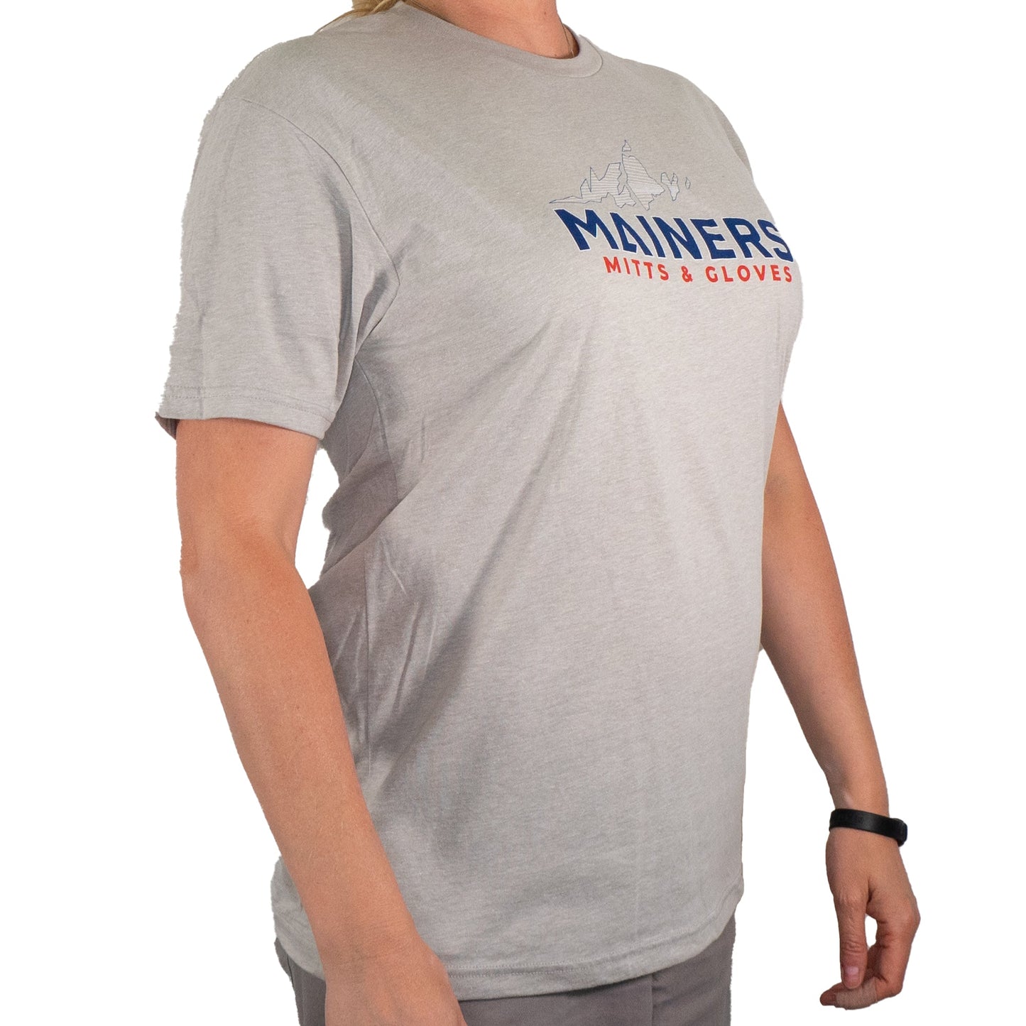 A person clad in a premium Mainers MG Tee, featuring the "Mainers" logo, stands with arms at their sides, poised for outdoor adventure.