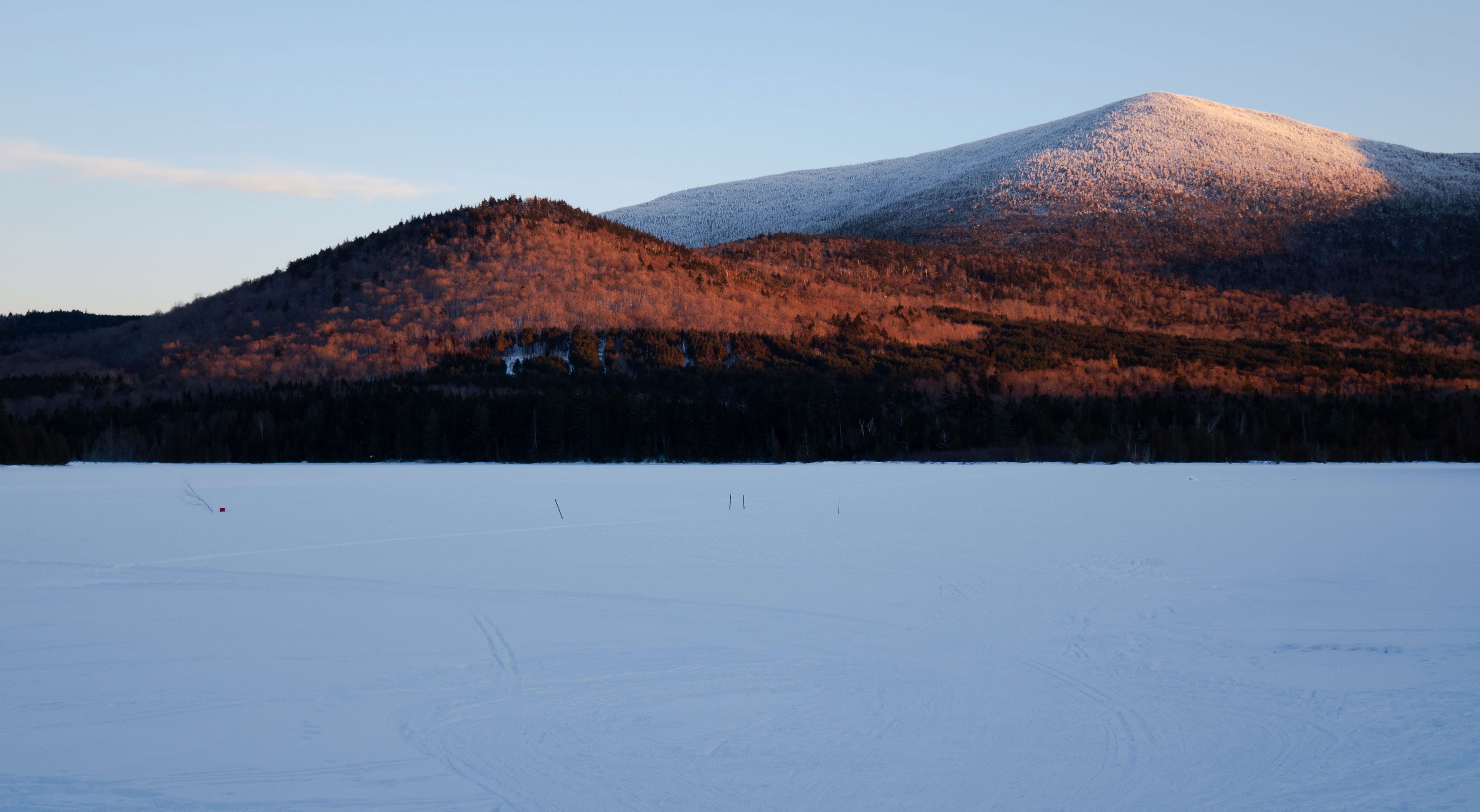A snowy mountain range under a clear blue sky with sunlight casting a warm glow on the peaks. The foreground features a frozen, snow-covered field.