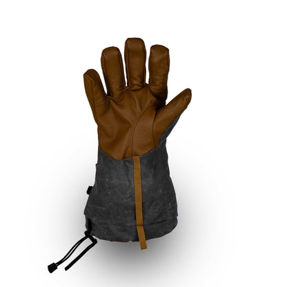 The Mainers Bethel Glove, designed for outdoor adventures, showcases a brown and black color scheme with a robust leather palm and extended cuff. Set upright against a simple white background, its rugged charm is elevated by the durable waxed canvas finish.