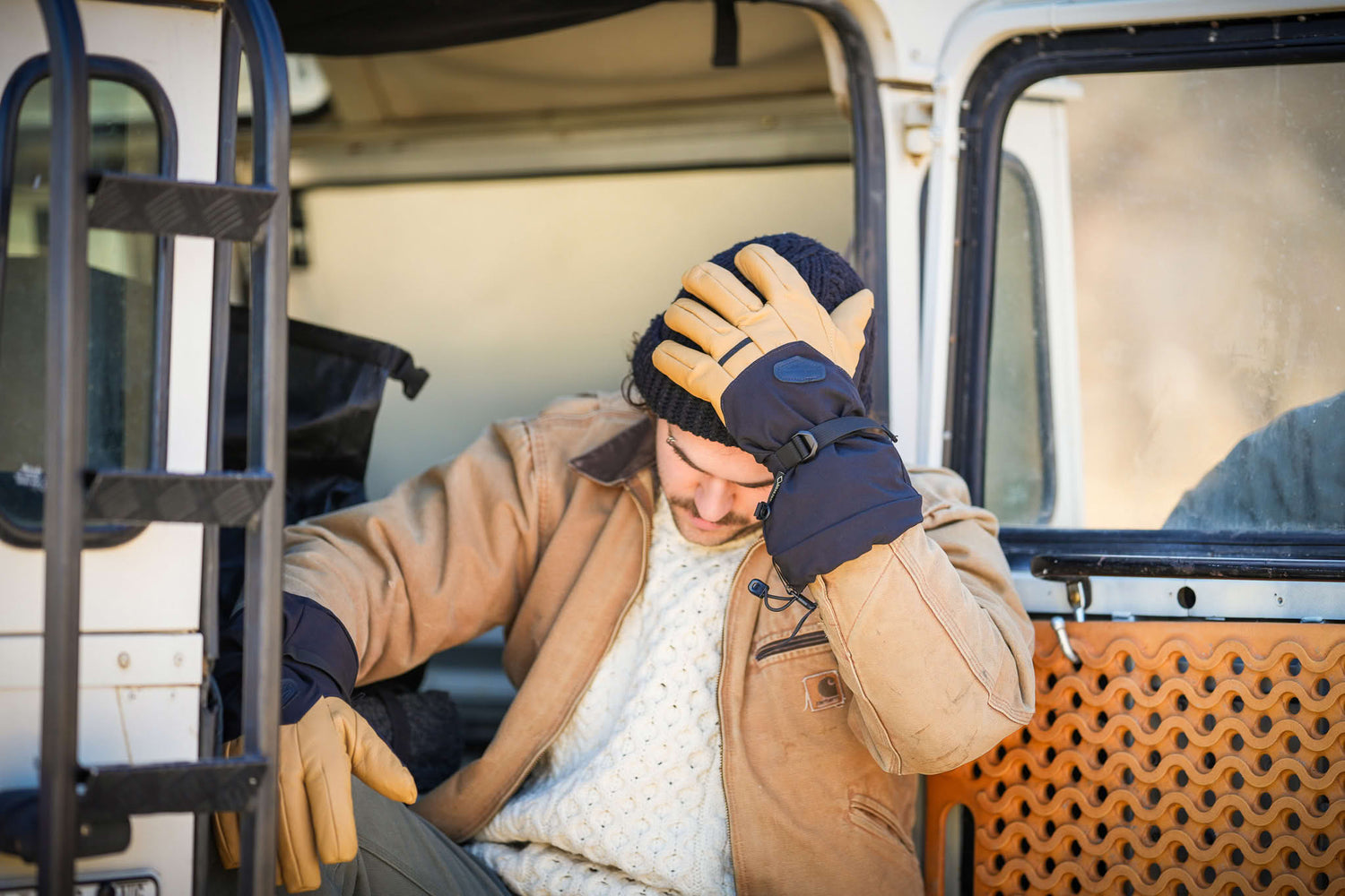 Man sitting in a vehicle, wearing a brown coat, knit hat, and yellow gloves, holds his head with one hand.