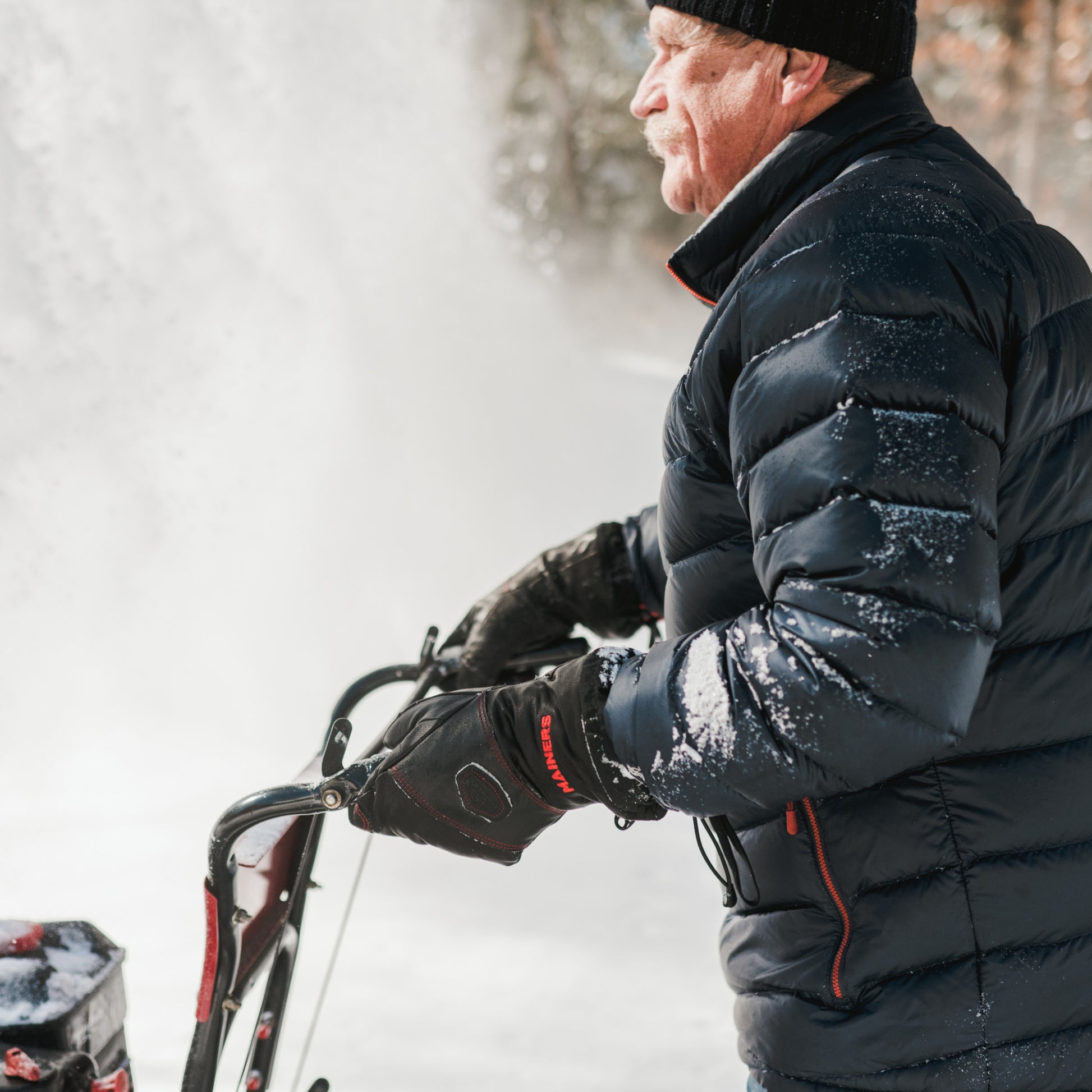 A man in a black leather coat and hat using a Mainers All-Leather Mitts snow blower in cold weather.